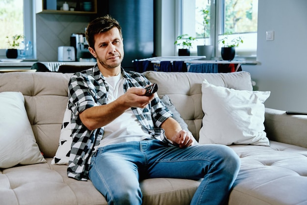 소파에 앉아 TV를 보고 있는 남자는 리모콘을 사용하여 채널을 전환하고 집에서 주말 여가를 즐기고 있습니다.