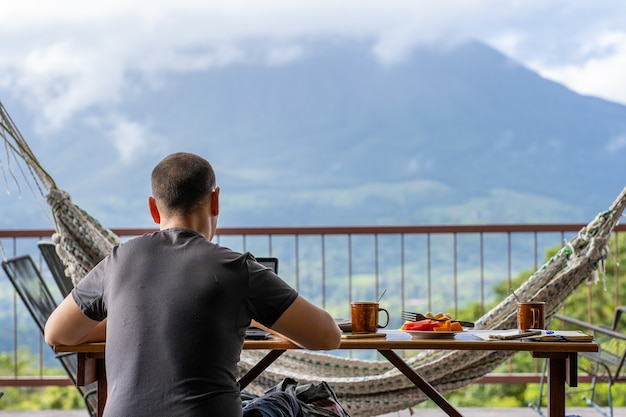 Uomo seduto su un tavolo con la colazione mentre si lavora con la vista di un vulcano di fronte a lui. costa rica