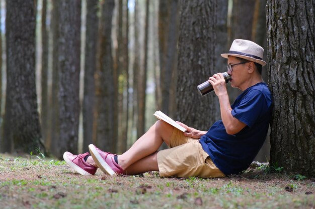 Photo man sitting under a tree in the pine forest drinking cold drink from a tumbler