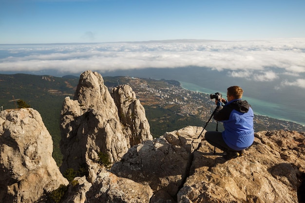 Man sitting with a tripod and photo camera on a high mountain peak above clouds city and sea