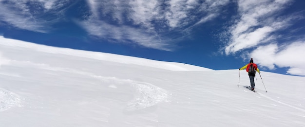 Man ski touring climbing snow-covered mountain under blue and cloudy sky