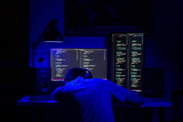 A man sleep at a computer in a room at a table at night with blue lighting and programs