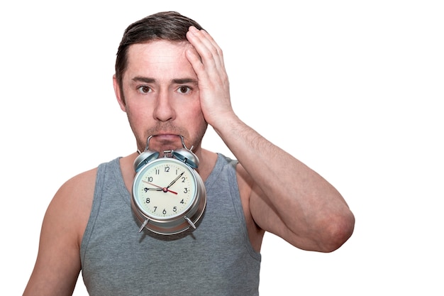 The man slept for work. A man holds an alarm clock in his teeth. Puzzled facial expression. Holds his head with his hand. Isolated white background.