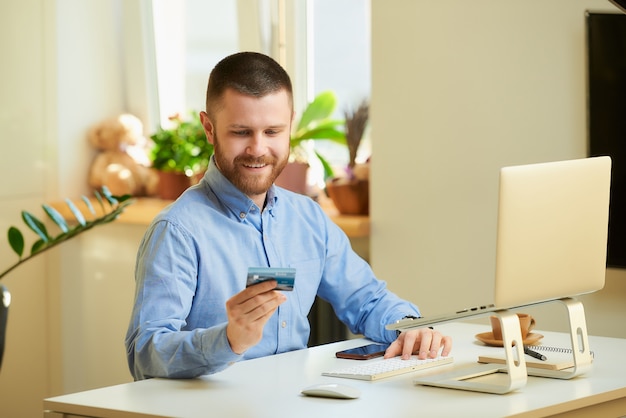 Un uomo che sorride e che esamina la sua carta di credito davanti al computer portatile a casa.