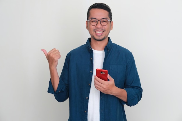 A man smiling and pointing to the right side with his thumb while holding mobile phone