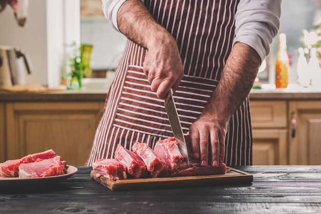 Man snijdt vers stuk rundvlees op een houten snijplank in de huiskeuken. Een man in een gestreept schort met een groot mes in zijn handen