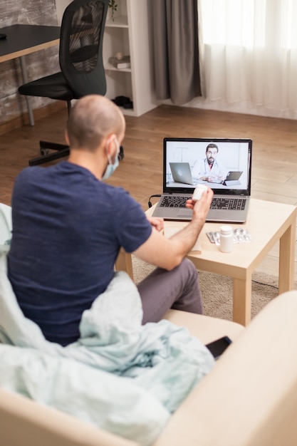 Man on sofa in a video call with doctor during quarantine holding pills bottle.