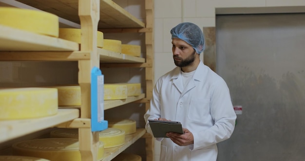 Specialista uomo in uniforme bianca che controlla la qualità dei formaggi lavorando nella fattoria di produzione contadino maschio presso il proprio caseificio