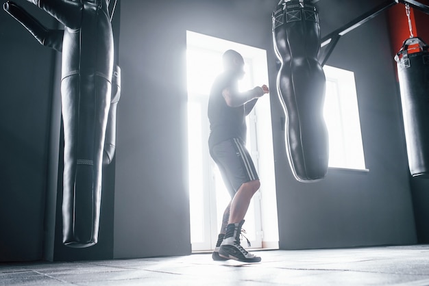 Man in sportive clothes training boxing in the gym with pushing bags. Illuminated by light.