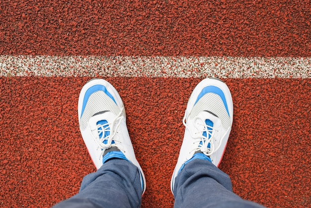 Man in sports white running shoes stands on the red jogging\
track in the stadium outside, close-up. top view, first person\
view