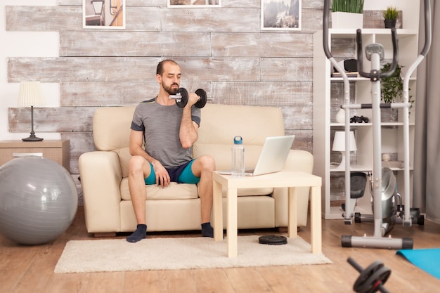 Uomo in abbigliamento sportivo con manubri che guarda l'allenamento online durante la pandemia globale.