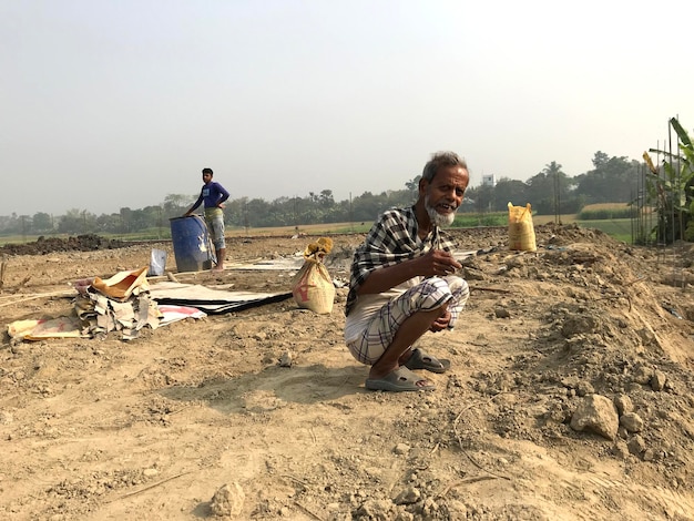 A man squats in a field with a bag of sand in the background.