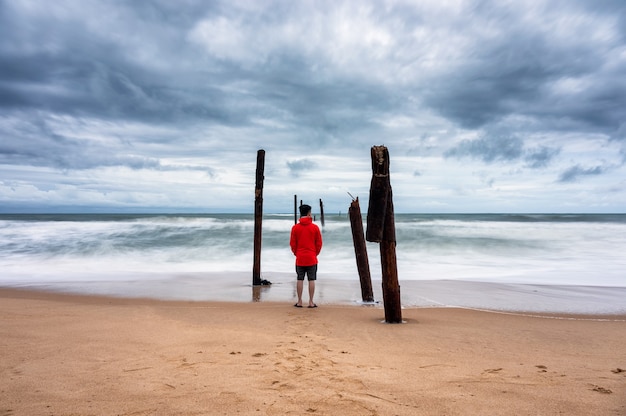 Man standing on the beach and big wave hitting decay wooden bridge in stormy weather at Pilai Beach, Phang Nga