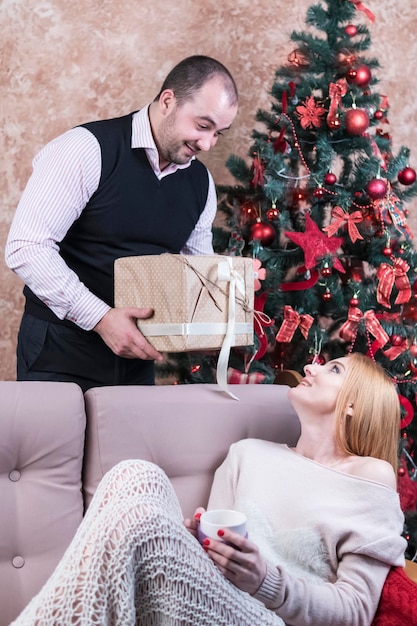 Man standing at the head of a woman gives her a Christmas present, against the background of the Christmas tree