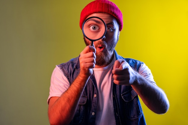 Man standing holding magnifying glass pointing to camera with\
surprised face