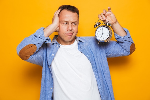 Man standing isolated over yellow wall holding alarm clock.