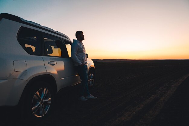 Человек, стоящий рядом с белой машиной, смотрит на закат.