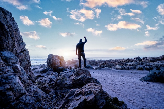 человек, стоящий на скале, вид сзади с поднятой рукой, глядя на закат над океаном