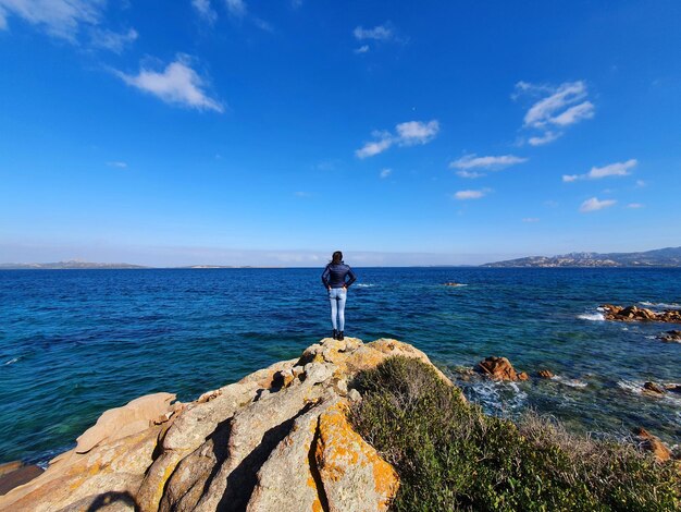 Man standing on rock by sea against blue sky