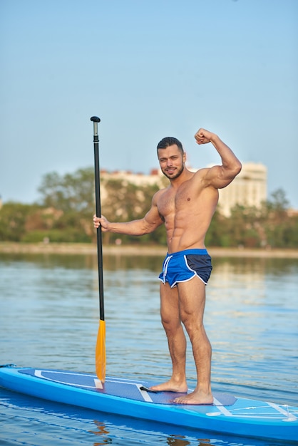 Man standing on sup board and showing biceps, holding long oar.