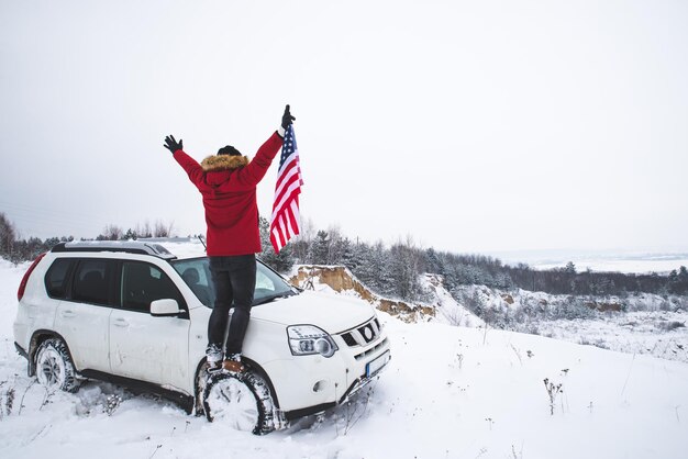 Uomo in piedi sulla cima della collina con la bandiera degli stati uniti nelle sue mani vicino a un'auto suv in cerca di una bellissima vista invernale