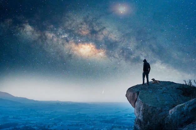 Man standing on the top of the mountain back view with his dog and Milky Way over the valley