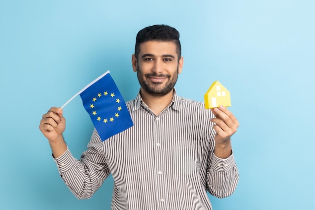 Uomo in piedi con casa di carta e bandiera dell'immigrazione dell'unione europea e acquisto di una casa in europa