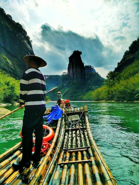 Photo man standing on wooden raft against rock cliffs and clouds