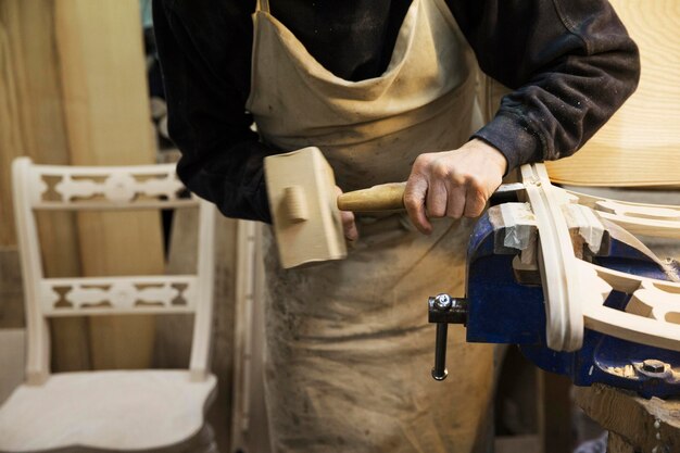 Foto uomo in piedi su un banco di lavoro in un laboratorio di falegnameria che lavora su una sedia di legno con un martello di legno e uno scalpello