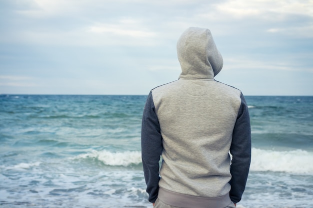 男はフードと海を見てトラックスーツのビーチの上に立つ