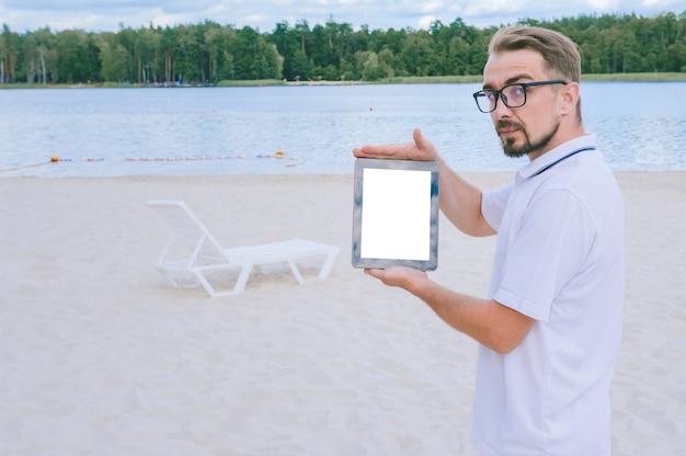 한 남자가 손에 모의 태블릿을 들고 해변에 서 있습니다. 갑판 의자와 물과 숲이 있는 모래를 배경으로.