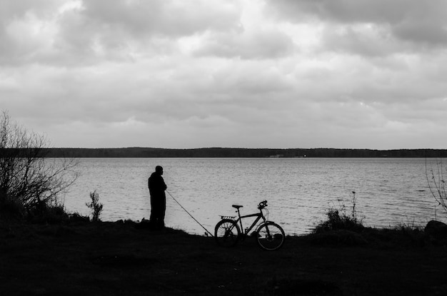 A man stands next to a bike and a bicycle on the shore of a lake.