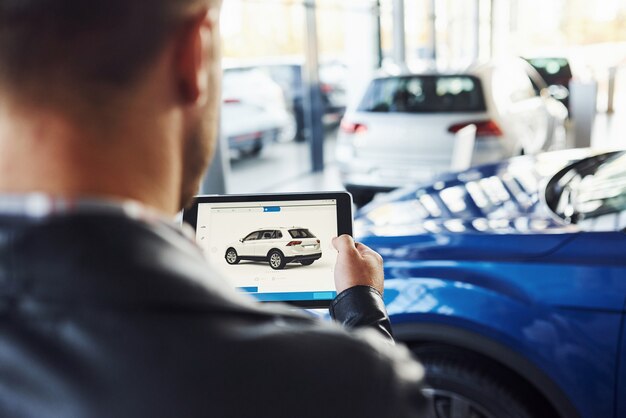 Man stands inside car salon with tablet in hands and looks at the vehicle picture.