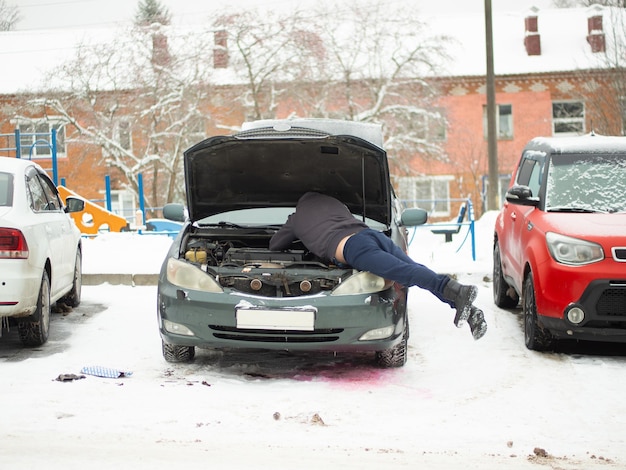 男は駐車場で冬の季節に現代の車の修理の開いたボンネットの下に立つ 男は寒い冬の車の手入れで車を始動しようとする