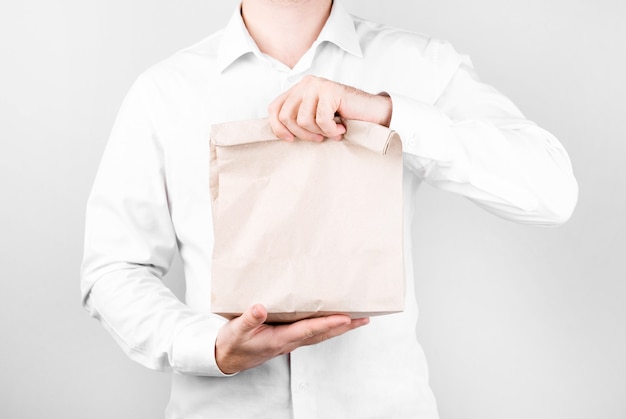 A man stands on a white wall in a shirt and with two hands holds out a paper bag instead of a plastic one, recycling, shopping and ecology concept