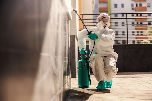 Man in sterile protective uniform crouching and spraying wall with disinfectant in order to prevent corona virus from spreading.