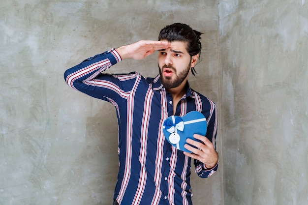 Man in striped shirt holding a blue heart shape gift box and looking forward to find the nominee