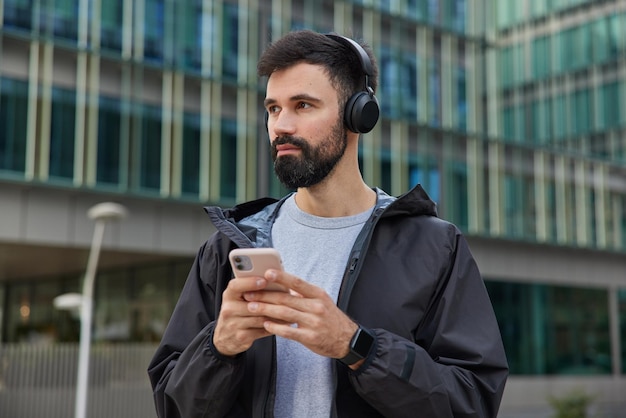 man strolls in city with modern gadget listens favorite music concentrated away poses on modern building downloads song to playlist uses media application