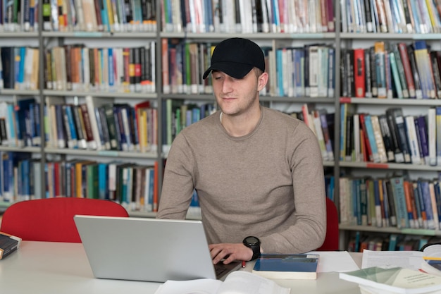Студент-мужчина в кепке работает над ноутбуком и книгами в школьной библиотеке
