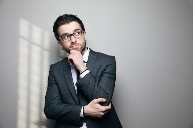 A man in a suit and glasses thinks and touches his chin on a gray wall