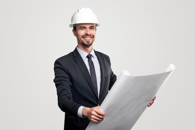 Man in suit and hardhat smiling and looking at camera while reading blueprint during work on architectural project against gray background