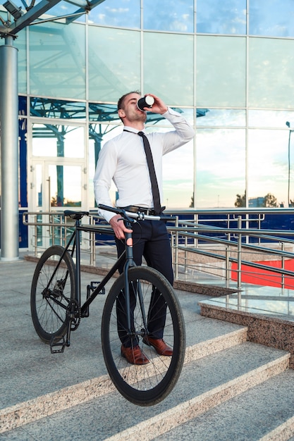 Man in suit holding a cup of coffee walking by the street with his bicycle.