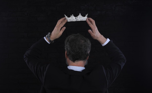 A man in a suit holds a crown over his head in front of a black background.