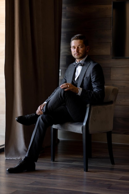 A man in a suit sits in a chair in front of a window.