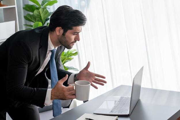 A man in a suit sits at a desk and talks to a laptop.