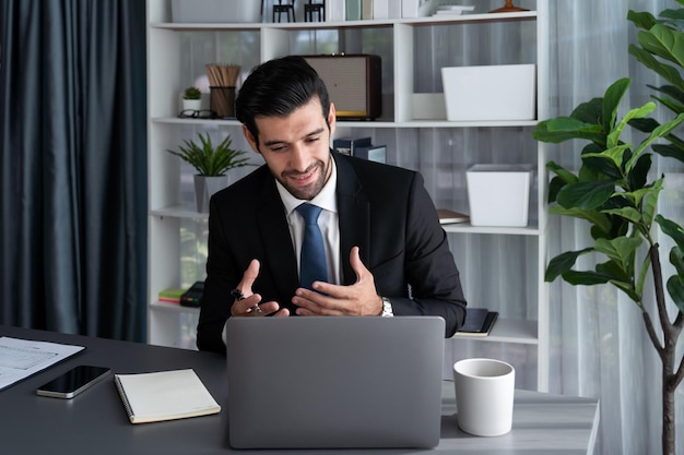 A man in a suit sits at a desk with a laptop and a coffee mug.