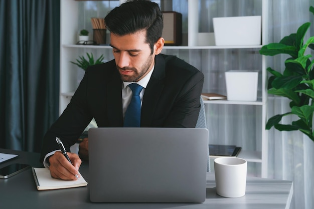 A man in a suit sits at a desk with a laptop and a pen.