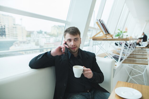 Un uomo in giacca e cravatta seduto a un tavolo in un luminoso caffè vicino alla finestra e parlando al telefono.