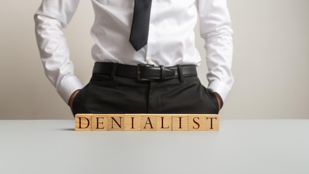 Man in a suit standing behind an office desk with a word Denialist spelled with wooden cubes in a conceptual image