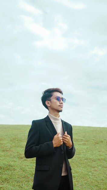 A man in a suit stands in a field with a cloudy sky behind him.